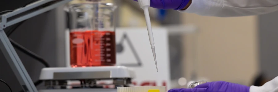 Gloved hand holding a pipette above a small container, with a beaker of red liquid on a lab plate in the background in the QAQC Lab on the University of Montana campus in Missoula, MT.