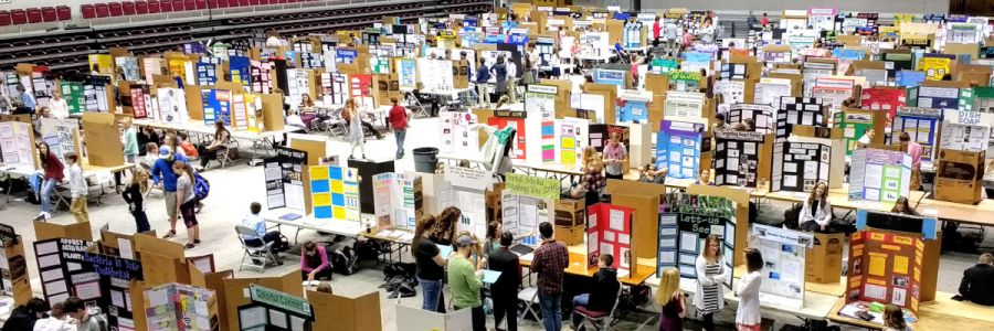 gym floor full of tables with science projects on each and people browsing and talking amongst them