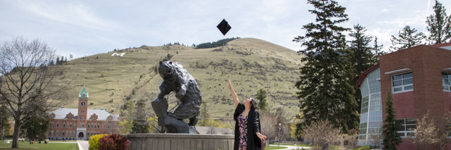 student throwing their graduation cap into the air in front of the Grizzly statue on the UM campus with University Hall, Mount Sentinel and the Payne Family Native American Center in the background