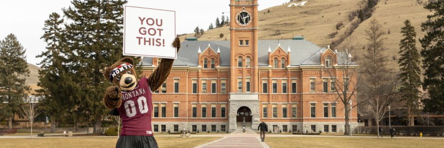 UM mascot Monte pointing at the camera and holding up a You Got This sign on the UM oval in front of University Hall