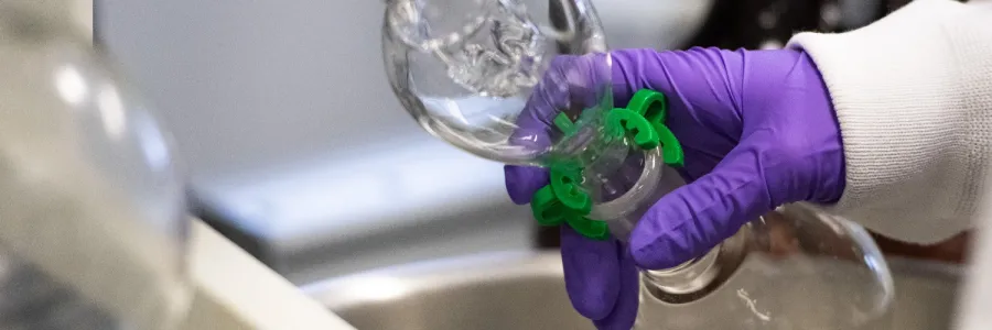 A UM doctoral candidate conducting research in a lab on the �����ؿ� campus with a gloved hand holding laboratory glassware with a green clamp over a lab sink..