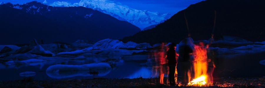group of people standing around a campfire at night with ocean glaciers and steep snowy mountains in the background