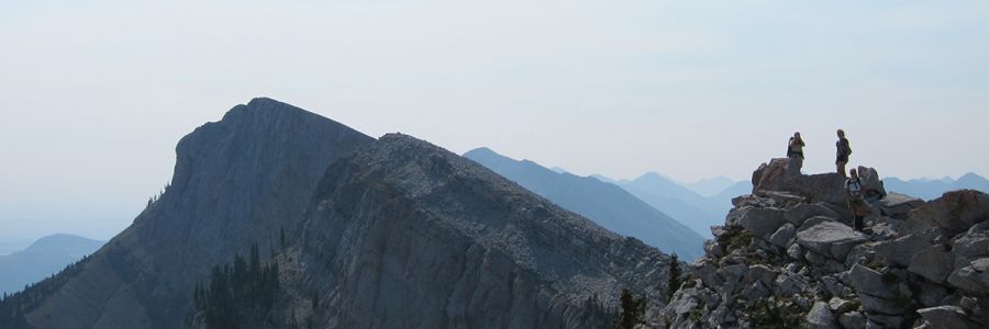 series of steep rocky peaks with three people standing on the top of one in the foreground