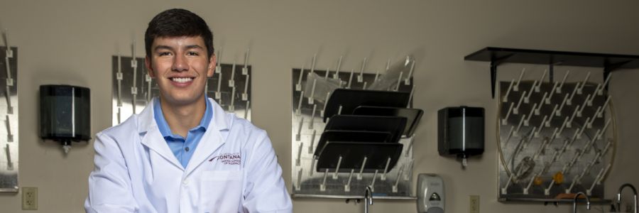 headshot of a student smiling in a lab coat with drying racks hanging on the wall behind them