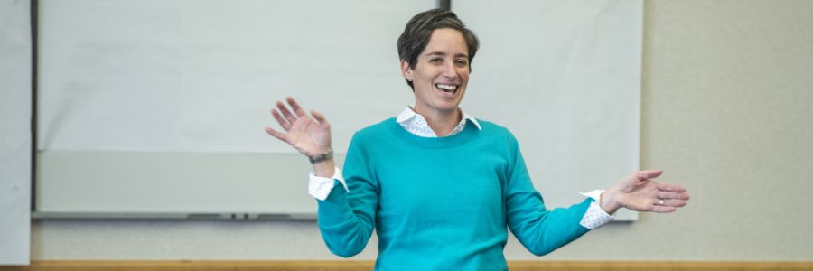 professor smiling and gesturing in front of a white board in a classroom