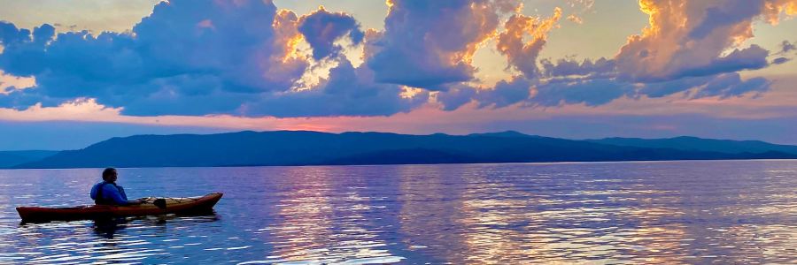 person in a kayak on a calm lake with dramatic sky and mountains behind