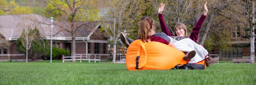 two students sitting on a bean bag together on the UM oval with one raising their hands and smiling