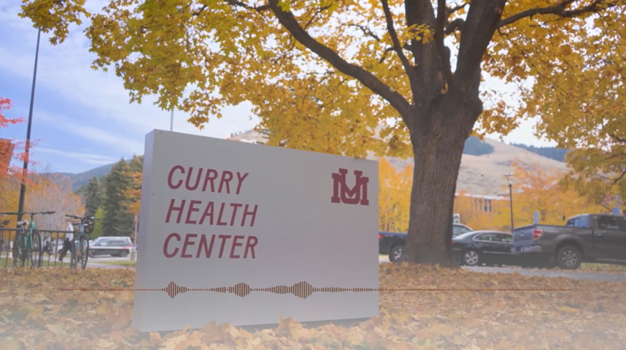 Photo shows gray sign that reads, 'Curry Health Center,' that's located in front of the building on Eddy Ave. at the University of Montana. Orange leaves and fall colors frame the sign.