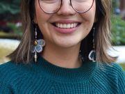 A female student with short brown hair, glasses, wearing a teal shirt, and dragonfly earrings.