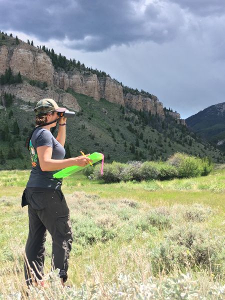 A woman stands looking through a scope toward a large body of water.