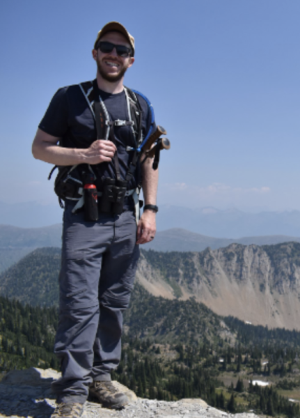 Matthew Trentman stands on top of a rocky mountain, smiling.