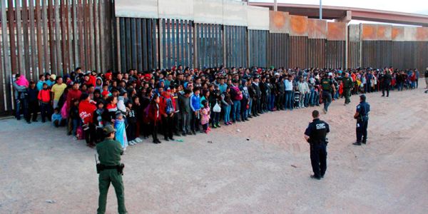 Hundreds of people in front of a rusty wall with border officers facing them