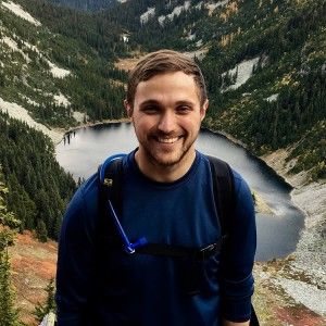 A person stands wearing a water bladder, and in the background there is a lake far below them.