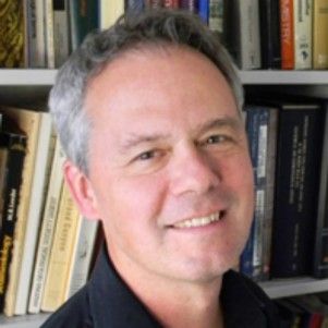 A person stands in front of a bookcase and smiles gently at the camera.