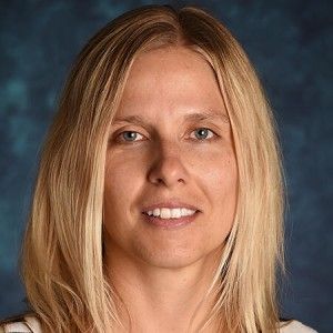 A person with blond shoulder length hair smiles at the camera in front of a blue background.