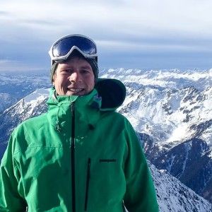 A person wearing heavy winter gear stands in front of a wide vista of snow capped mountains.