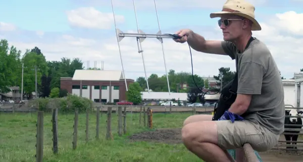 A research crouches in a field, holding a radio tranceiver.