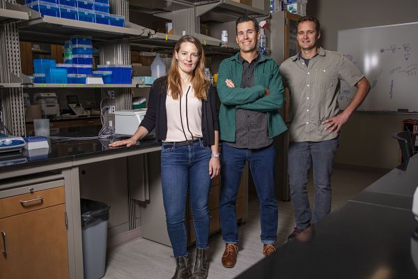 Three researchers stand in front of a lab bench, smiling at the camera