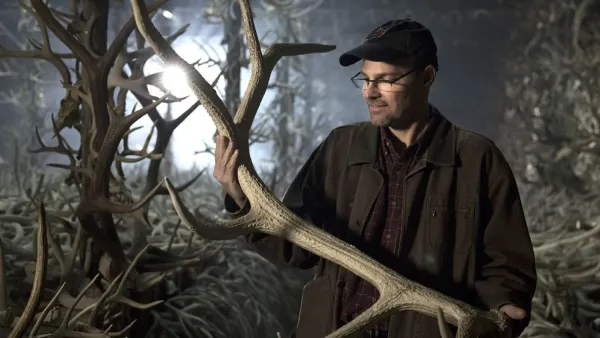 A person stands in front of the camera, holding a massive elk antler