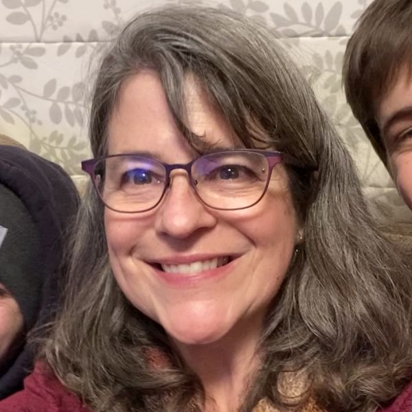 A person with shoulder-length hair and glasses smiles at camera.