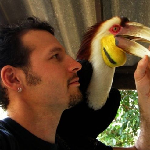 A person with short black hair and a goatee look to the right with a toucan perched behind them.