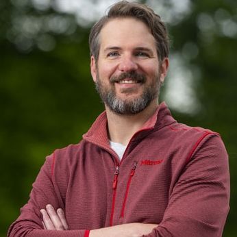 A person with a salt and pepper beard cross their arms and smiles at the camera.