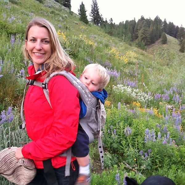 A person with long blond hair hikes with a baby bjorn on their back.