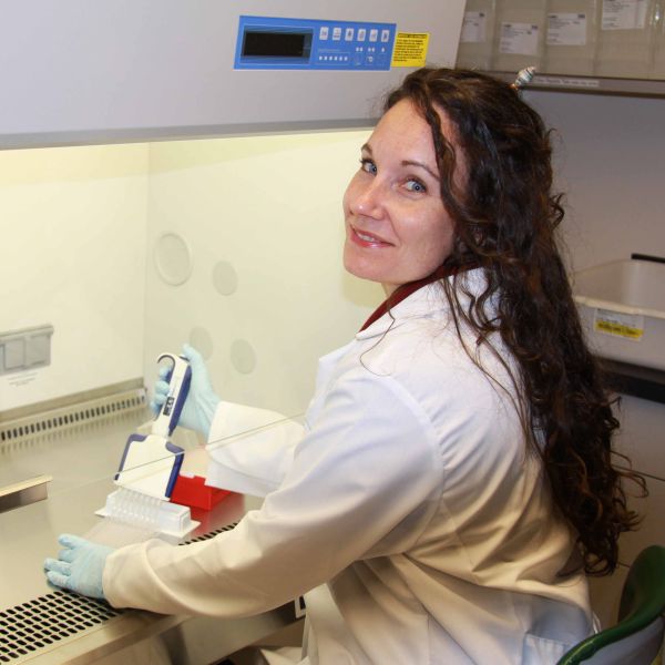 A person with long brown hair wears a labcoat and sits in front of a biosafety cabinet.