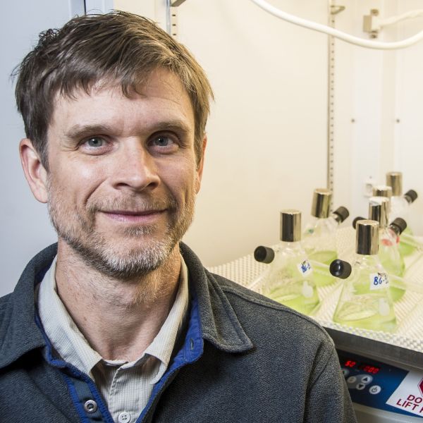 A man with short light brown hair stands in front of an incubator and grins at the camera.