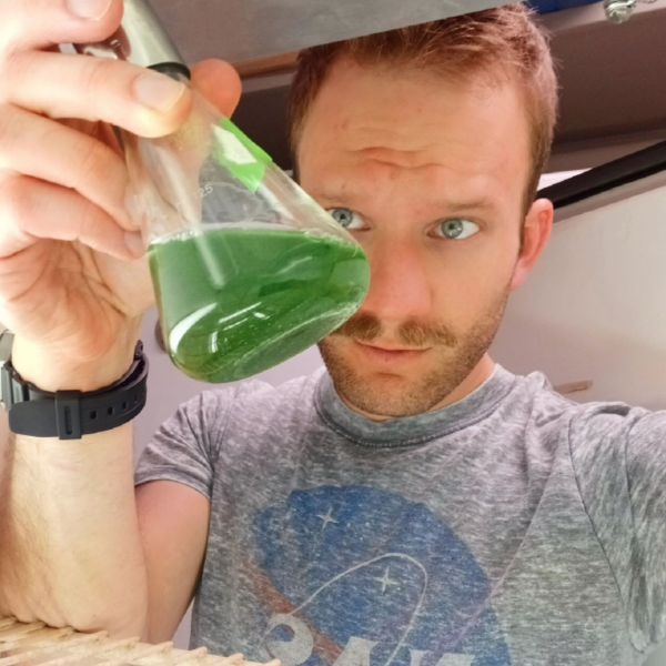 A person with short blond hair and moustache looks seriously at a flask of green liquid.