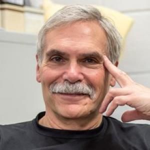 A person with gray hair and moustache smiles brightly at the camera.