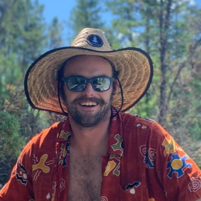 A person with a wide brim straw hat and a colorful shirt smile at the camera.