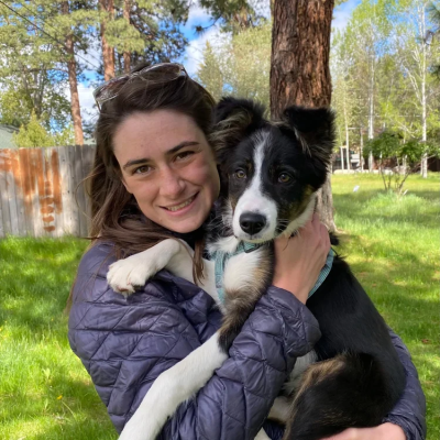 A person wearing a purple puffy jacket hugs a puppy and smiles at the camera.
