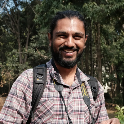 A person holding a camera smiles at the camera standing in front of a lush forest.