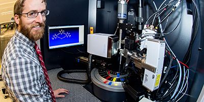 A researcher smiles next to a sophisticated scientific instrument used for molecular analysis, with a computer screen displaying a molecular structure