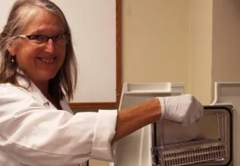 A lab technician wearing a white coat and gloves is smiling while retrieving a sample from a laboratory freezer