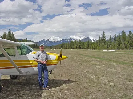 Erick Greene stands beside a light aircraft on a grassy airstrip with snow-capped mountains in the background.