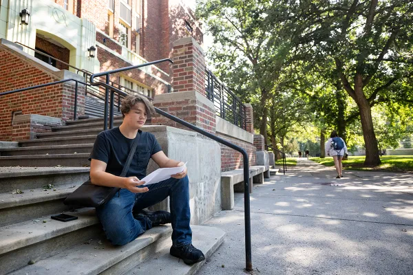 A student looks over a course syllabus while sitting on the steps of the Fine Arts Building
