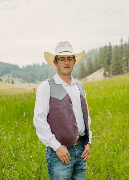 Chase Kellett in Western wear stands posed with his thumbs in his pockets in a sunny field.