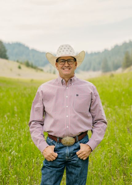 Coach Kory Mytty in Western wear stands posed with his thumbs in his pockets in a sunny field.