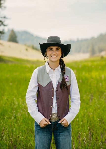 Hailey Gliko in Western wear stands posed with her hands on her vest in a sunny field.
