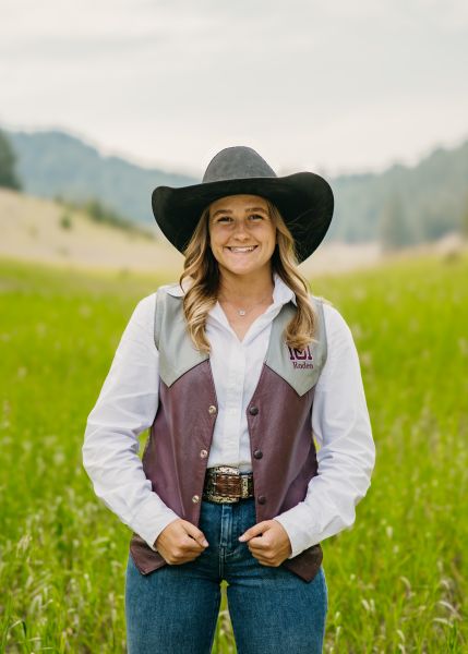 Kyra Pietrass in Western wear stands posed with her hands on her vest in a sunny field.
