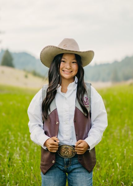 Lexi Murer in Western wear stands posed with her hands on her vest in a sunny field.