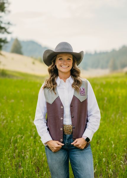 Maryah Winn in Western wear stands posed with her hands on her vest in a sunny field.