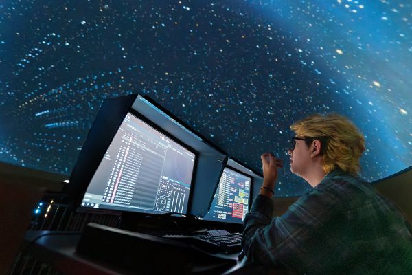 planetarium presenter sits at control panel with night sky in the background