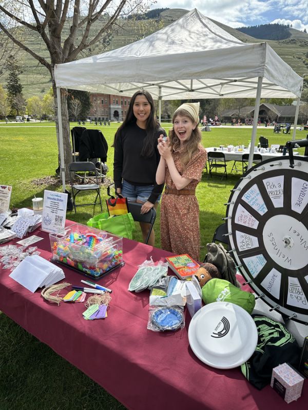 Two young students stand at a table filled with colorful items and a spinning prize wheel in the stress less booth during an event on the UM oval. One woman is smiling excitedly while the other poses with a bag.