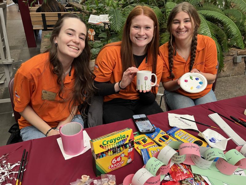Three volunteers in orange shirts smile while showcasing their decorated mugs and art supplies at an event in the University Center