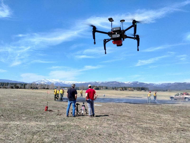 A flying drone in the foreground against a blue sky with a fire team working in the background in a field