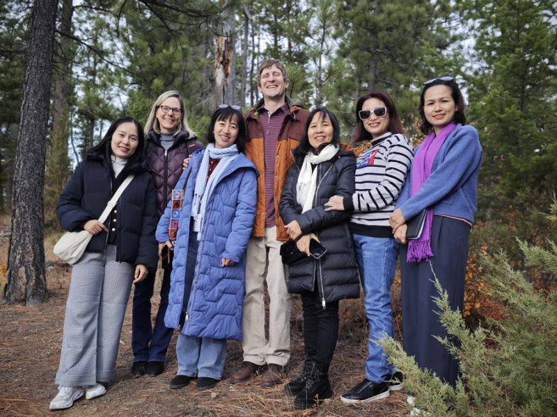 Group of eight people posing together in a forest setting, surrounded by tall pine trees