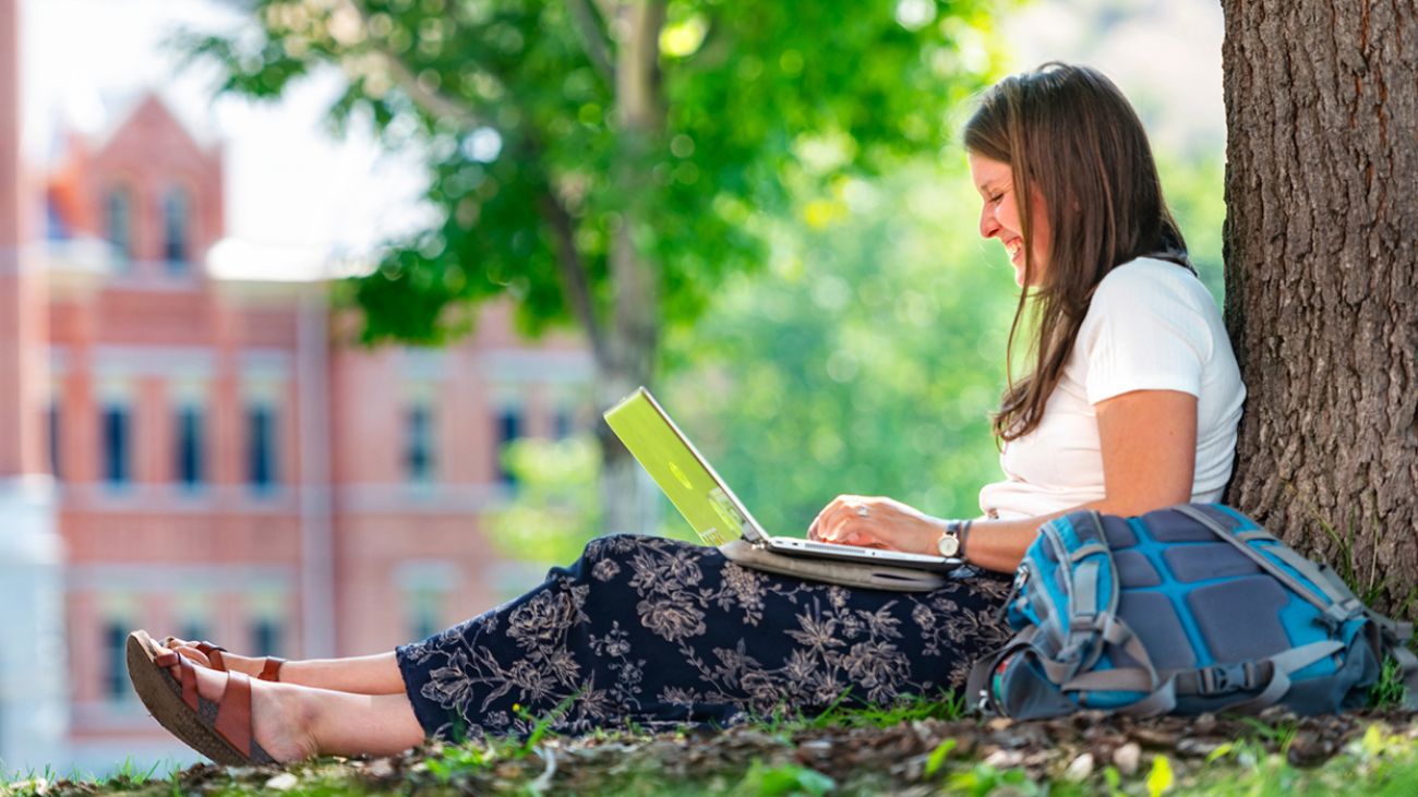A student smiles while working on her laptop as she sits again a tree near Main Hall